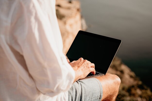 Digitaler Nomadenmann mit Hut sitzt ein Geschäftsmann mit Laptop auf den Felsen am Meer bei Sonnenuntergang