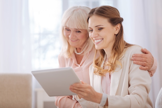 Digitales Gerät. Erfreute positive junge Frau, die lächelt und auf den Tablettbildschirm schaut, während sie zusammen mit ihrer Mutter sitzt