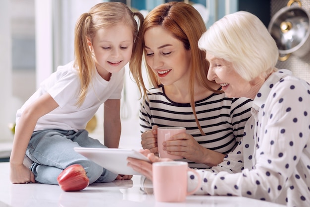 Digitalización contemporánea. Niña linda sentada en la encimera de la cocina y viendo un video en la computadora portátil junto con su madre y su abuela tomando café