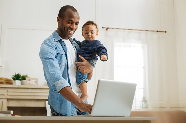 Digitando. atraente alerta jovem pai afro-americano segurando seu filho pequeno e digitando no laptop enquanto está à mesa e uma lareira ao fundo