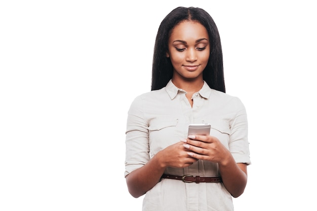 Digitando mensagem para um amigo. Mulher africana jovem confiante segurando um telefone celular e olhando para ele em pé contra um fundo branco