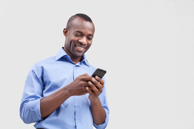 Digitando uma mensagem. Homem negro alegre digitando algo no celular e sorrindo enquanto fica isolado no cinza