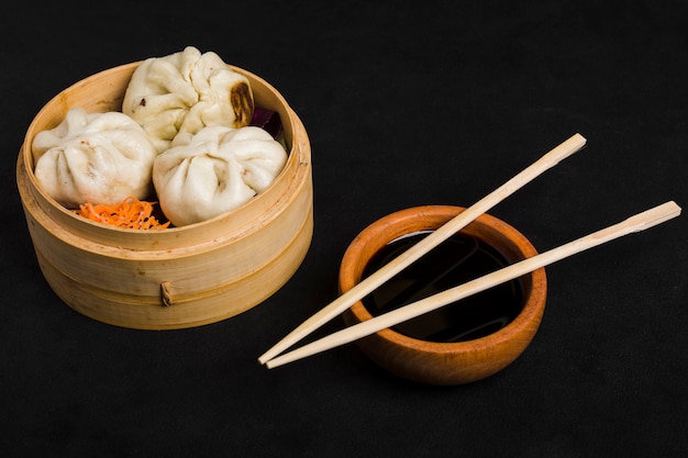 Foto dim sum junto con la ensalada de zanahoria servida en una cesta pequeña de vapor y un tazón de salsa de soja con palillos sobre fondo negro