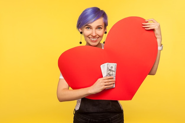 Dinero y amor. Retrato de una chica hipster sonriente y positiva con el pelo corto violeta en overoles de mezclilla con un gran corazón de papel y un montón de dólares. aislado sobre fondo amarillo, tiro de estudio interior