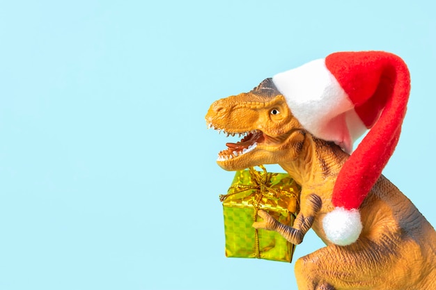 Dinosaur Rex en rojo sombrero de Santa Claus tiene caja de regalo dorada en sus patas sobre fondo azul.