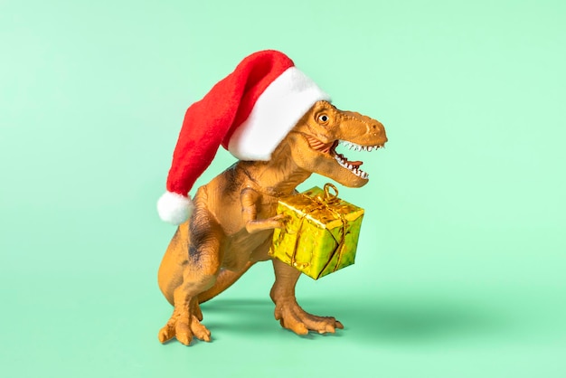 Dinosaurier Rex in rotem Weihnachtsmann-Hut hält goldene Geschenkbox in seinen Pfoten auf blauem Hintergrund