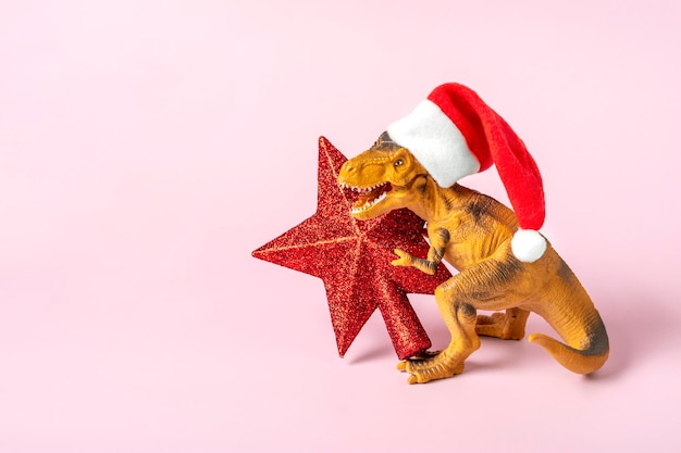 Dinosaurio Rex con sombrero rojo de Santa Claus sostiene una estrella en sus patas sobre fondo rosa