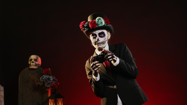 Diosa espeluznante de la muerte jugando videojuegos en línea, sosteniendo el controlador para jugar competencia en la consola. Mujer con traje de santo dios de los muertos para celebrar la cultura mexicana. Disparo de mano.