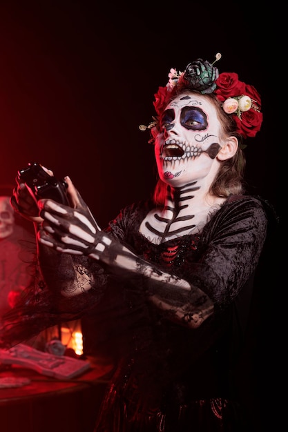 Diosa de la muerte jugando videojuegos en línea con joystick en la consola, con maquillaje de calavera y traje tradicional de santa muerte. mujer disfrutando de la competencia de juegos en el día de los muertos.