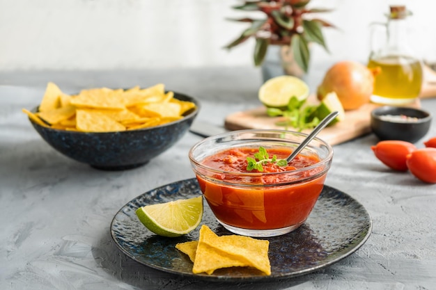 Foto dip de lima picante con tomate con tortilla de nachos en un tazón y los ingredientes principales a un lado. fondo gris