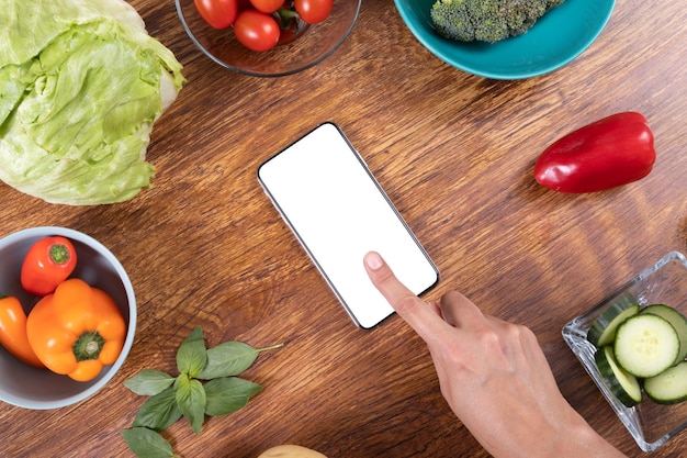 Directamente encima de una foto de una mano recortada usando un teléfono inteligente en blanco en medio de varios alimentos en la mesa, espacio para copiar