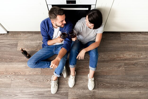 Foto directamente encima de la vista de la hermosa pareja positiva en jeans sentado en el piso de parquet en la cocina y bebiendo vino tinto en aniversario