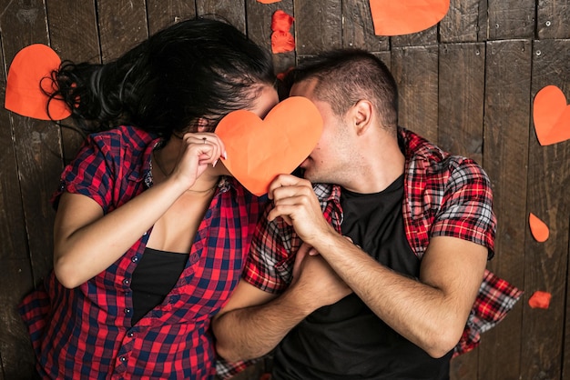 Foto directamente encima de la vista de una pareja joven y amorosa que cubre sus rostros con un trozo de papel en forma de corazón y se besan mientras están acostados en el piso vacaciones del día de san valentín