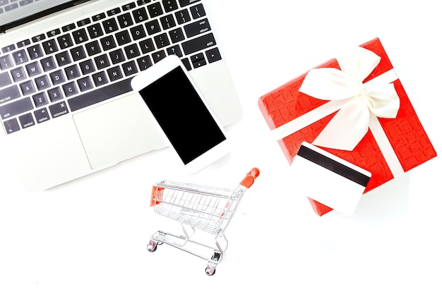 Direkt über dem Laptop befindet sich ein Miniatur-Einkaufswagen und eine Geschenkkiste mit Kreditkarte auf weißem Hintergrund
