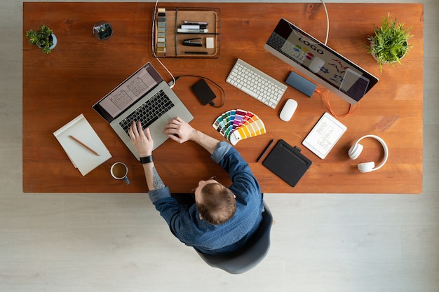 Direkt über der Ansicht des Mannes mit Tätowierung, die am Holztisch sitzt und UI-Entwurf auf Laptop bearbeitet