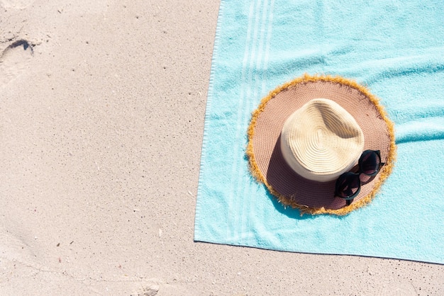 Diretamente acima da foto de óculos de sol com chapéu e toalha azul na praia durante o dia ensolarado. Inalterado, verão, proteção, ausência, conceito de férias.