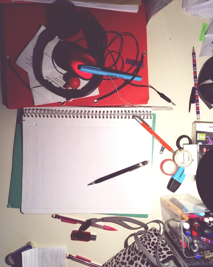 Foto diretamente acima da foto do livro e canetas com fones de ouvido na mesa