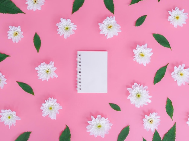 Diretamente acima da vista do caderno em branco com flores brancas e folhas verdes em fundo rosa