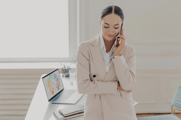 Diretora ocupada em traje formal tendo conversa de telefone celular falando no telefone e discutindo novo projeto com co-parceiro Empresária trabalhando no escritório leve com laptop na mesa de madeira