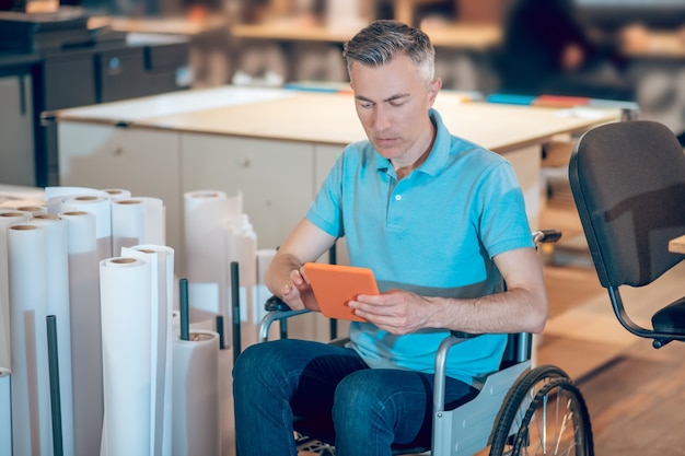 Discapacidad, empleo. Hombre discapacitado enfocado atento sentado en silla de ruedas trabajando con tableta en espacio de oficina