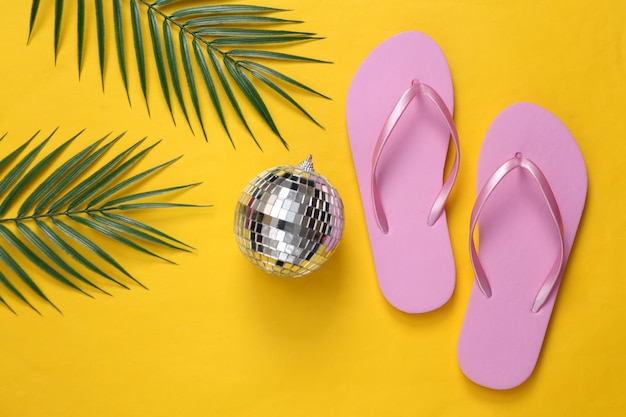 Disco-Kugel mit Flip-Flops und Palmblättern auf gelbem Hintergrund Draufsicht Flach liegend Minimalismus Strandparty und Ruhekonzept