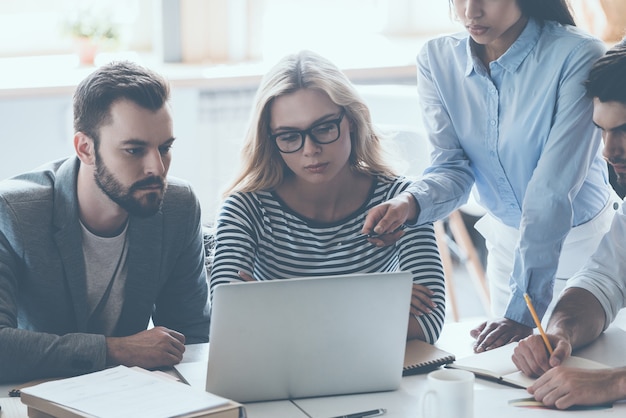 Discusión de negocios. Grupo de jóvenes empresarios sentados en el escritorio de la oficina y discutiendo algo mientras miran juntos la computadora portátil