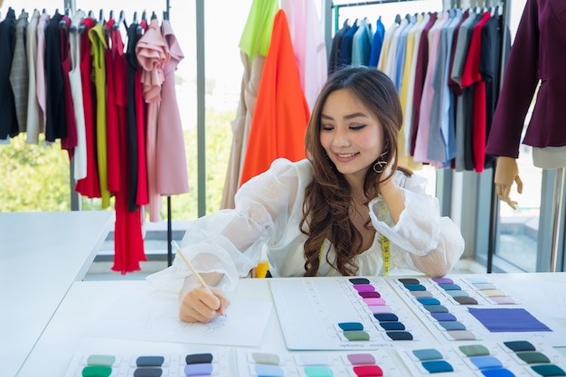 Diseñador asiático femenino lindo y enérgico que trabaja en el escritorio en la sala de diseño entre la ropa y la muestra del color de la tela.