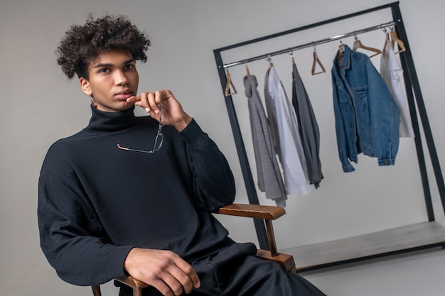 Foto diseñador. chico joven afroamericano vistiendo ropa negra sentado en una sala de exposición