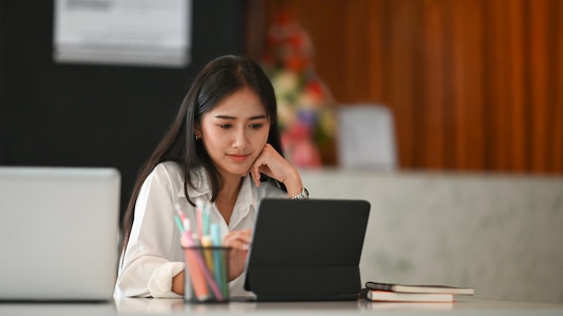 Diseñador gráfico atractivo de la mujer joven que trabaja en la oficina en casa y que usa la tableta de la computadora en el escritorio blanco.