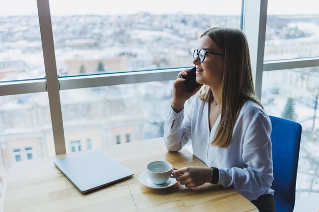 Diseñador gráfico que se conecta a Internet inalámbrico para crear archivos web llamando a un colega para discutir ideas mujer sonriente con anteojos que está hablando en un teléfono inteligente y tomando café en un café