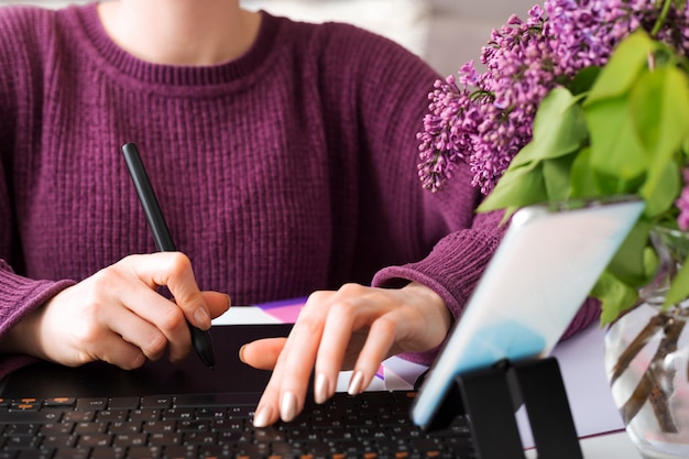 Diseñador gráfico retocador independiente trabaja desde casa. La mujer se basa en la tableta gráfica en la acogedora oficina en casa usando la computadora portátil, computadora. Trabajo remoto. Blogger grabando tutorial de transmisión en línea usando el teléfono