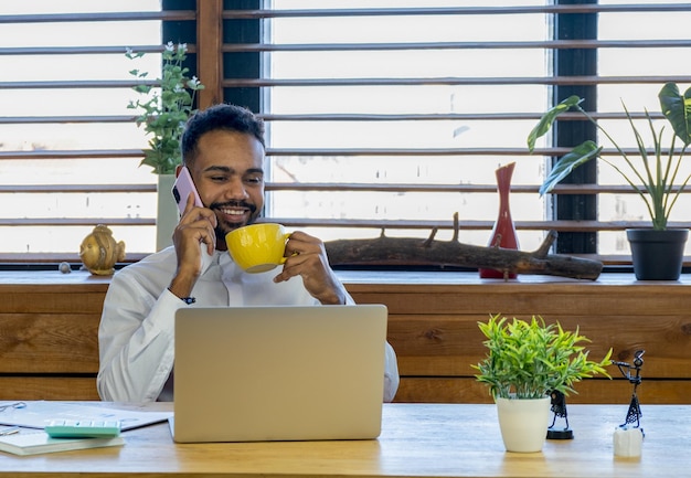 Diseñador masculino hablando por teléfono móvil y tomando café Hombre africano sentado en su escritorio y trabajando