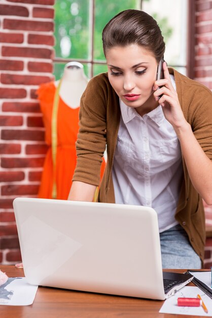 Diseñador de moda ocupado. Confiado diseñador de moda joven mirando la computadora portátil y hablando por el teléfono móvil mientras está sentada en su lugar de trabajo