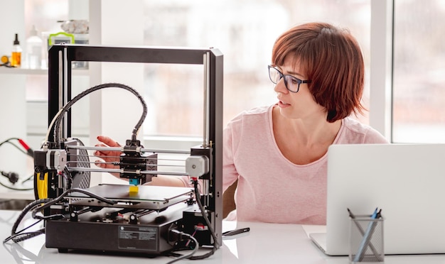 Diseñador de mujer con gafas trabajando con impresora 3D y portátil gris claro en proyecto