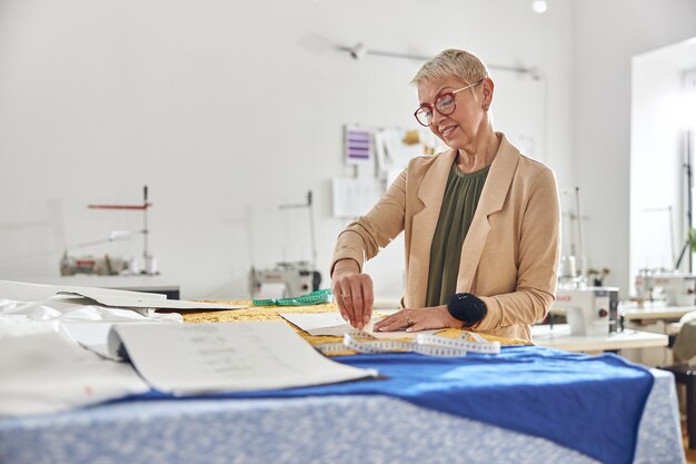 Diseñador de mujer madura dibuja un patrón en tela de color en una gran mesa de corte en el taller