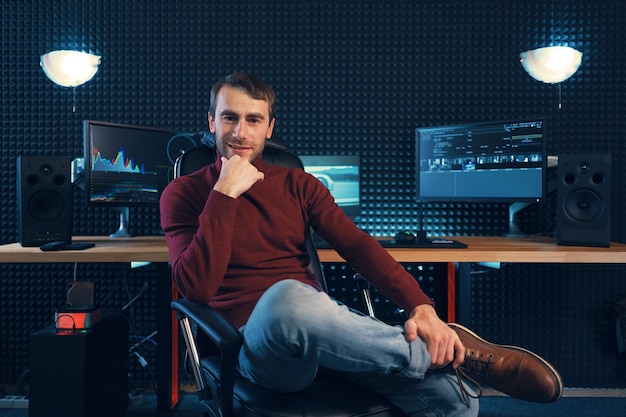Diseñador o editor exitoso sentado pierna sobre pierna en un sillón de cuero mirando a la cámara, con monitores y pared insonorizada en el fondo con espacio de copia