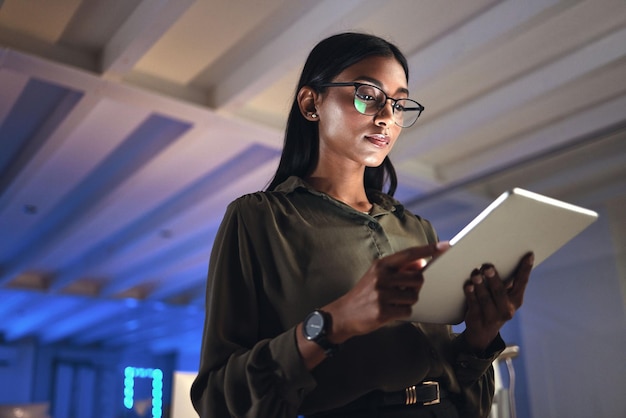 Diseñador de tabletas e investigación de mujeres serias en la oficina de inicio de negocios por la noche en la fecha límite Profesional creativo de tecnología o empresario indio que lee información por correo electrónico o aplicación en una empresa de neón