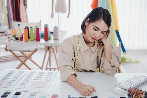 La diseñadora asiática está pensando y diseñando ropa para que los clientes pidan artículos en el escritorio del diseñador en el estudio.