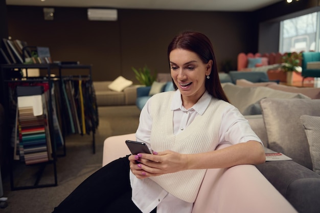 Diseñadora de interiores encantadora y elegante morena mujer caucásica de mediana edad vestida de forma informal escribiendo texto en su teléfono móvil sentada en un elegante sofá rosa moderno en una sala de exposición de una tienda de muebles