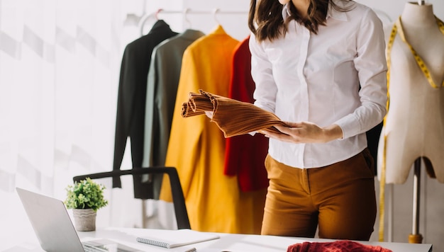 Diseñadora de moda l joven mujer asiática que trabaja con una tableta portátil y sonríe mientras está de pie en el taller