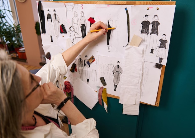 Diseñadora de moda de mujer madura dibujando ropa en la pared con dibujos anclados y bocetos de una nueva colección de ropa en un taller de sastrería