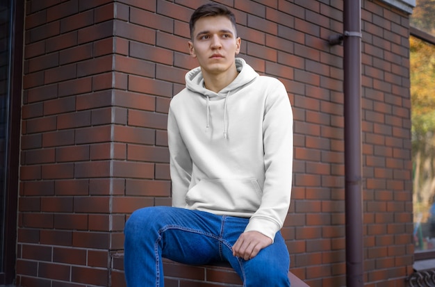 Diseño de una capucha blanca en un chico joven contra una pared de ladrillos, vista frontal. Mockup de ropa casual para presentación en su tienda. Plantilla de sudadera con capucha para hombre de moda