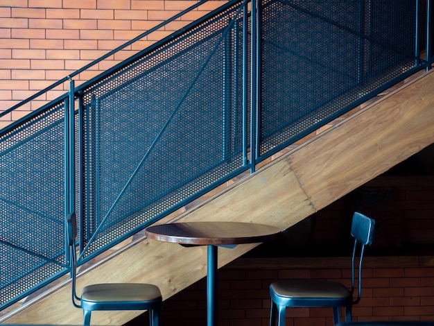 Diseño de decoración de café Mesa redonda de madera con dos taburetes de bar modernos cerca de escaleras de metal sobre fondo de pared de ladrillo