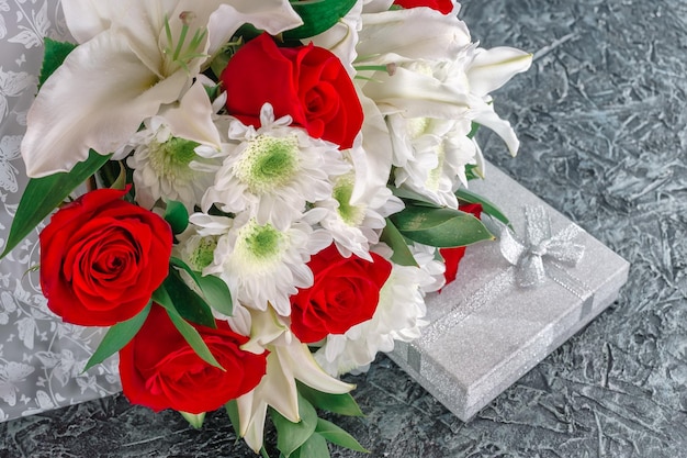 Diseño de fondo para felicitar a las mujeres Un ramo de flores de rosas, lirios y crisantemos con un regalo en una caja plateada