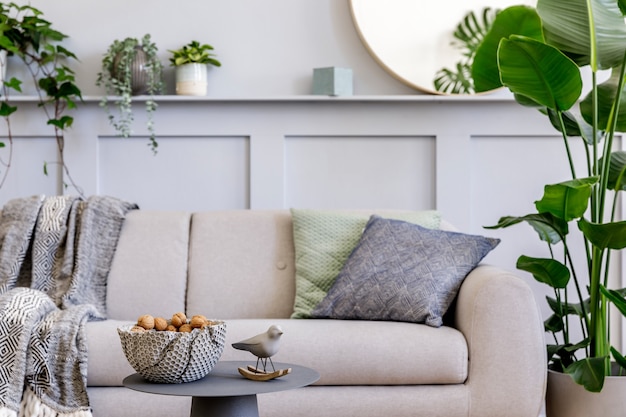 Diseño de interiores de sala de estar escandinava con elegante sofá gris, mesa de café, planta tropical, espejo, decoración, almohadas, cuadros y elegantes accesorios personales en la decoración del hogar moderno.