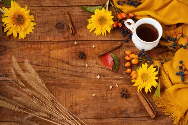 Diseño de otoño. Una taza de té se encuentra sobre una mesa de madera. Hermoso entorno con pañuelo amarillo, bayas y flores de girasol. Alrededor de las espiguillas de canela y las hojas de otoño. Copie el espacio. Endecha plana