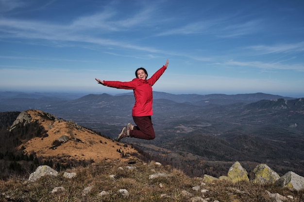 Disfruta de la aventura La viajera con chaqueta roja brillante salta alegremente en la cima de la montaña