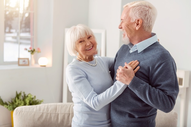 Disfruta bailando. Feliz anciana encantadora sonriendo mientras baila vals con su marido en la sala de estar