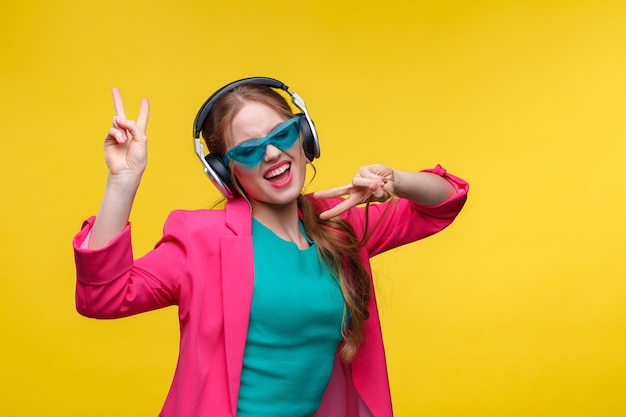 Disfruta escuchando música. Mujer joven pelirroja en auriculares escuchando música. Niña sonriente divertida en auriculares y chaqueta rosa bailando y cantando sobre fondo amarillo. Manejo de la relajación y el estrés.