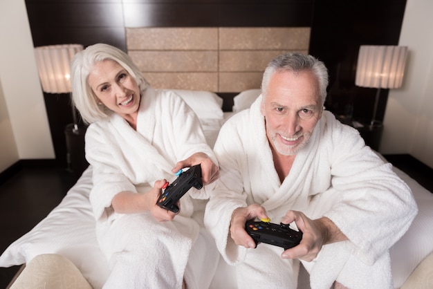 Disfrutamos de nuestro tiempo libre. Pareja de ancianos sonriente alegre sosteniendo consolas de juegos mientras está sentado en la cama y expresando alegría